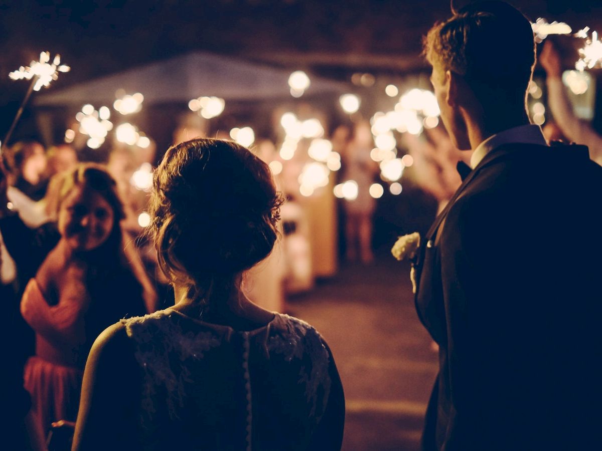 A couple stands with their backs to the camera, facing guests holding sparklers in a festive, dimly-lit setting, possibly a celebration or wedding.