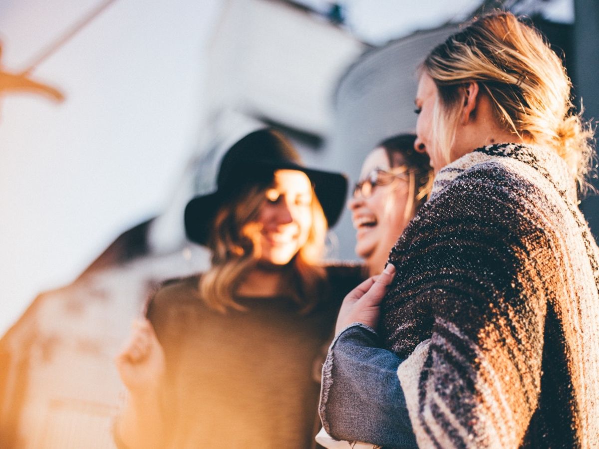 Three women are standing together, smiling and laughing, with one wrapped in a cozy blanket and another wearing a hat in soft, natural light.