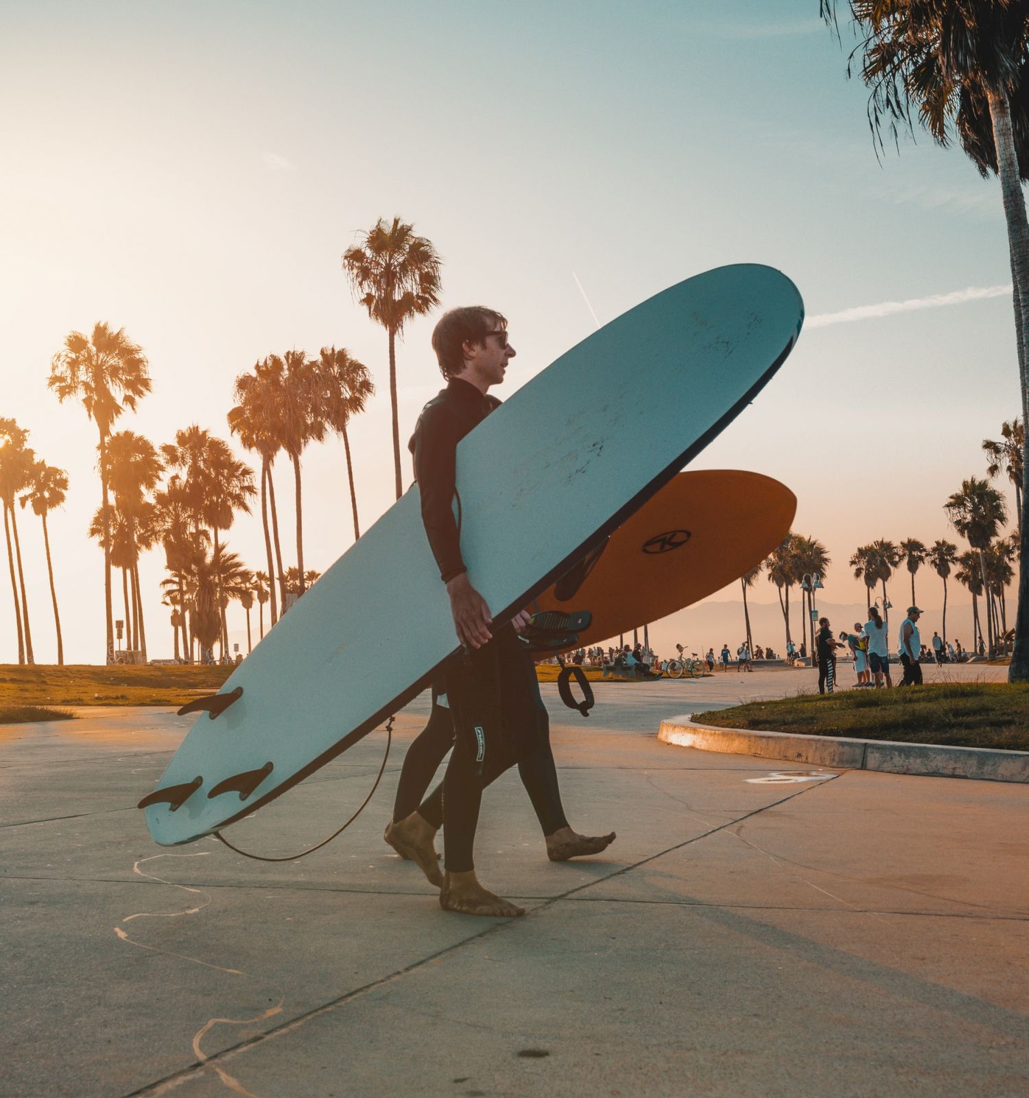 Two surfers carrying their surfboards walking along a palm tree-lined pathway at sunset, with clear skies and a warm glow.