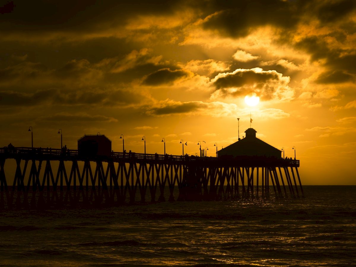 This image shows a pier extending into the ocean at sunset with golden clouds and a silhouetted structure at the end of the pier.