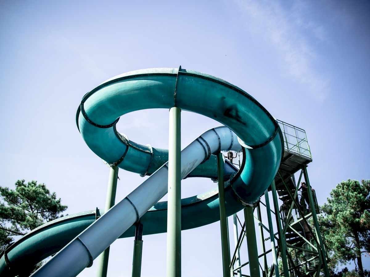 A large, spiraling water slide with blue tubes, supported by green beams, stands in an outdoor setting with trees in the background.