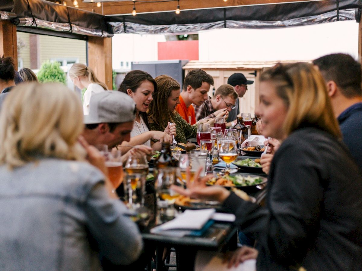 A group of people are gathered around a long table, enjoying food and drinks in an outdoor setting with festive lights overhead.