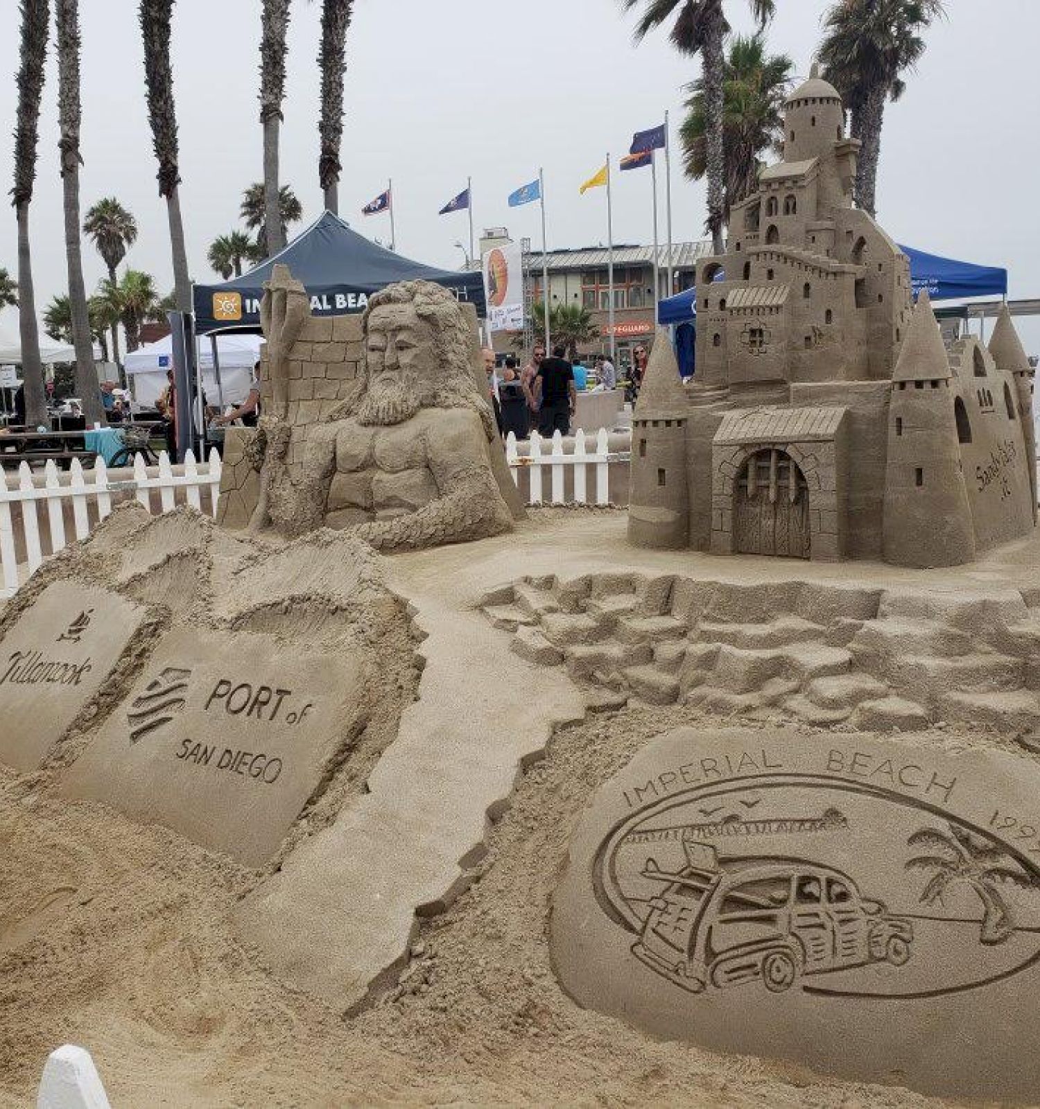 The image shows intricate sand sculptures, including a castle and a series of designs, at a beach event with people and palm trees in the background.