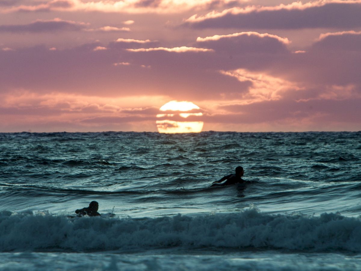 Two people are swimming in the ocean during a beautiful sunset, with the sun partially hidden behind clouds on the horizon.