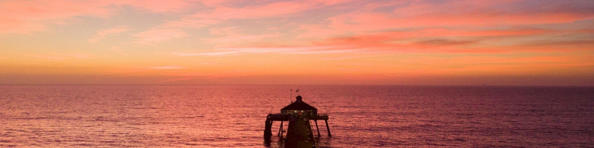 A long pier extends into a vast ocean, beneath a vibrant pink and orange sunset with a partly cloudy sky meeting the horizon line.