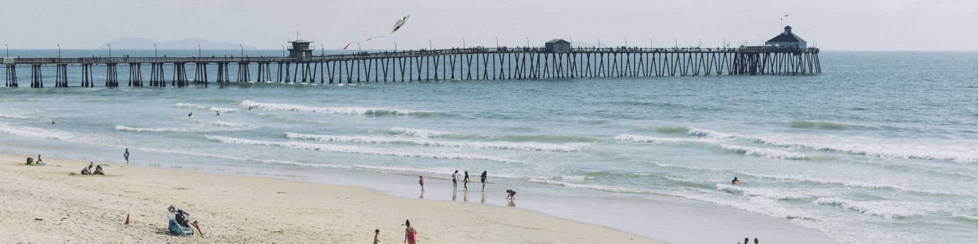 People are walking and relaxing on a sandy beach with a long pier extending into the ocean in the background, under a cloudy sky.