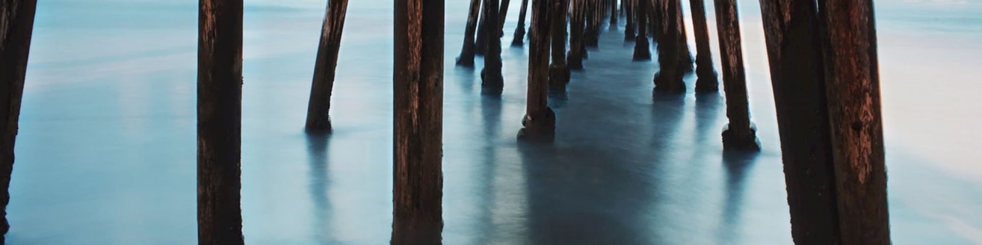 The image shows the view under a wooden pier, featuring numerous vertical beams extending into calm, reflecting water beneath a serene sky.