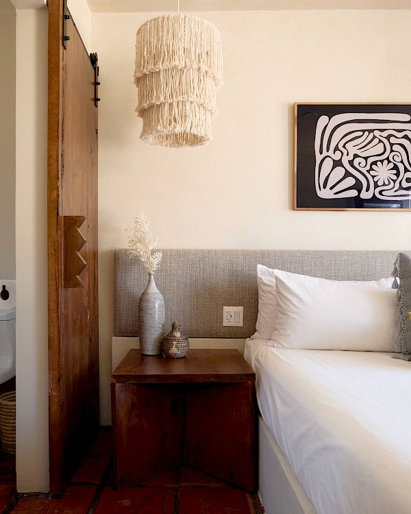 This image shows a cozy bedroom with a bed, wooden nightstand, vase, and decorative light fixture, with a sliding barn door partially open.