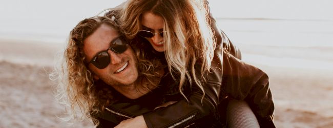 Two people wearing sunglasses are at the beach; one is giving the other a piggyback ride, both are smiling and seem to be enjoying the moment.