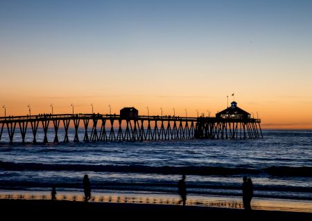 A picturesque pier extends into the ocean at sunset, with silhouetted figures on the beach and a vibrant sky in the background.