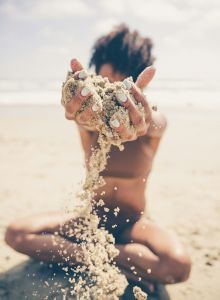 A person is sitting on a beach, holding up and letting sand fall through their hands, with the ocean visible in the background.