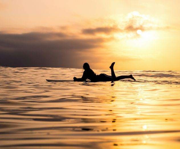 A person is lying on a surfboard in the water, silhouetted against a golden sunset with a calm sea and clouds in the background.