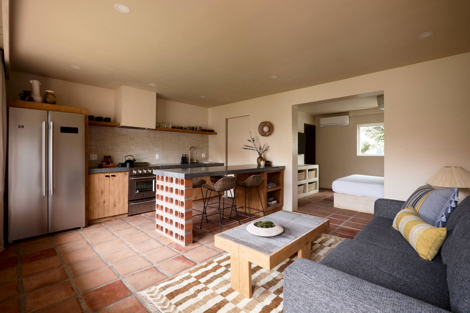 This image shows a cozy open-plan living space with a kitchen, dining area, and a lounge with a gray sofa. A bedroom is visible in the background.