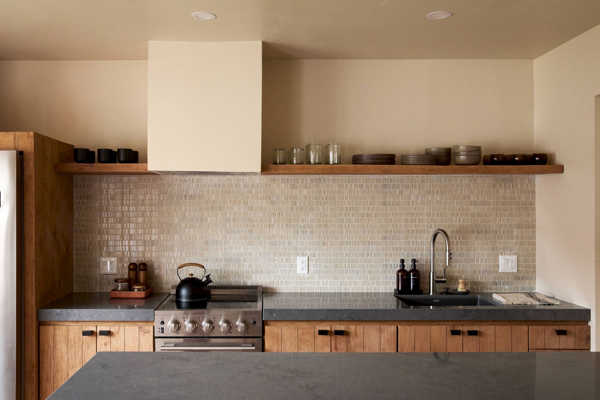 A modern kitchen with wooden cabinetry, a tiled backsplash, a stove with a kettle, a sink, and minimalistic decor on an upper shelf.