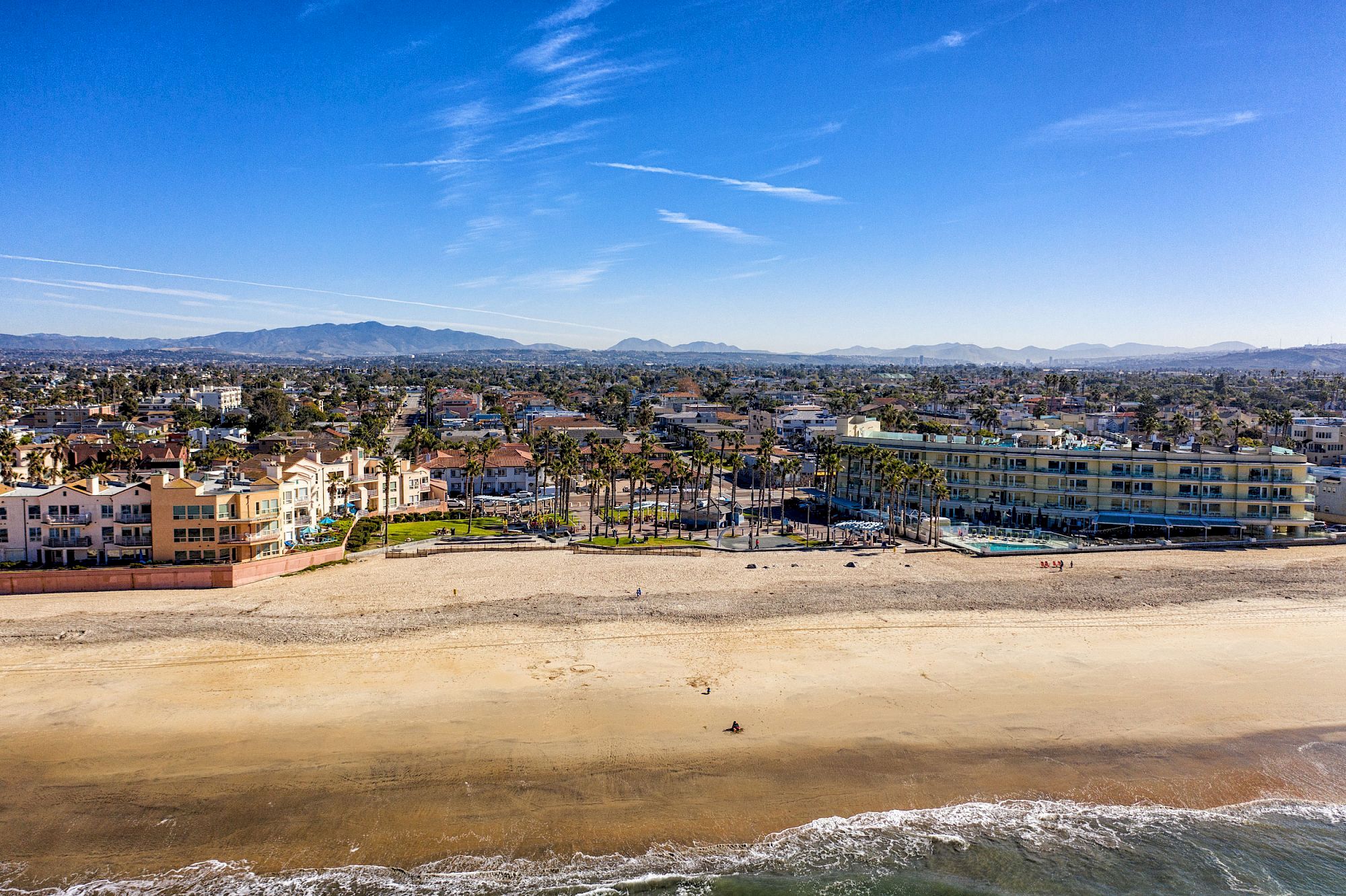 Aerial view of a coastal town with a sandy beach in the foreground, waves, buildings, palm trees, and mountains in the background.