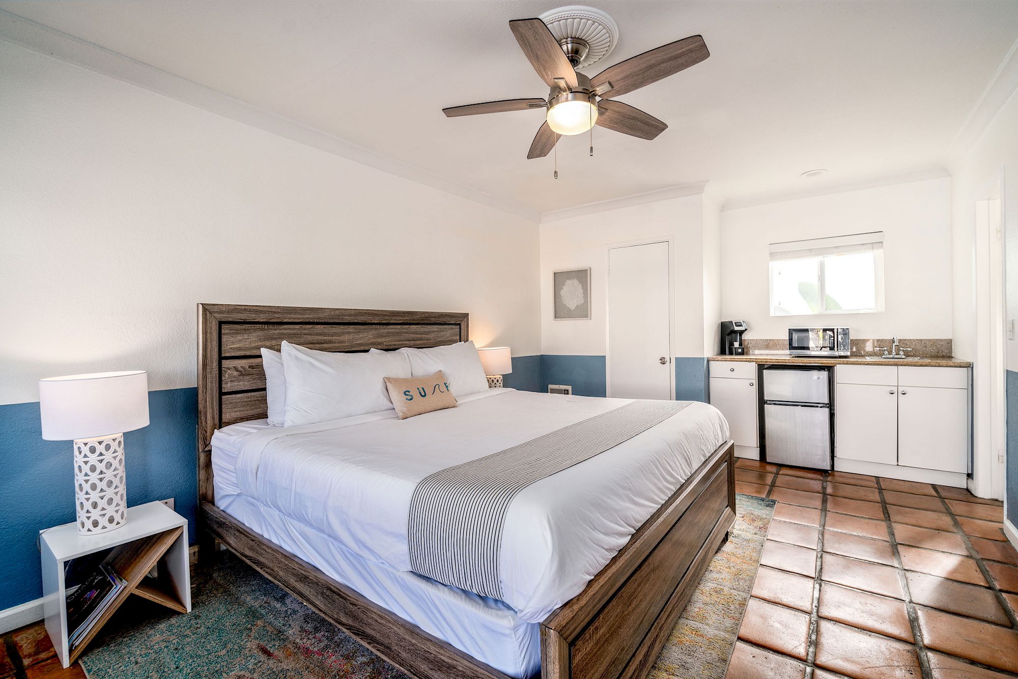 A cozy bedroom with a king-sized bed, ceiling fan, two nightstands with lamps, a kitchenette, and tiled floor. The bed has a decorative pillow.