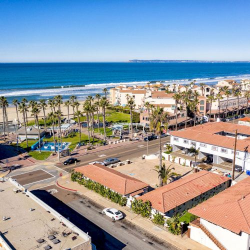 An aerial view of a coastal town with buildings, palm trees, a road, and a beach along the ocean with a clear blue sky in the background.