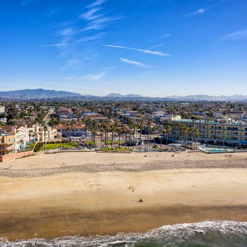 The image shows a beach with buildings and houses in the background, a few people walking along the sandy shore, and mountains in the distance.