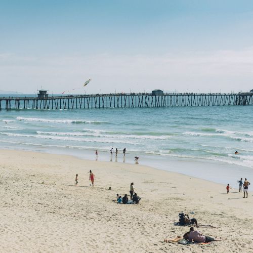 People walking and relaxing on a sandy beach with a long pier extending over the ocean in the background under a clear sky.