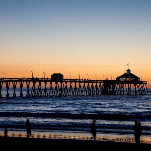 A pier extending into the ocean at sunset with silhouetted people walking on the beach and along the pier, as waves gently crash onto the shore.