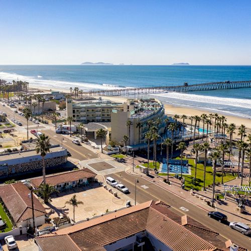 This image shows a coastal town with a long pier extending into the ocean, palm trees, buildings, roads, and a sunny beach along the shore.