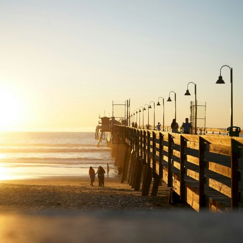 The image shows a pier extending into the sea during sunset, with two people walking on the beach and street lamps lining the pier.