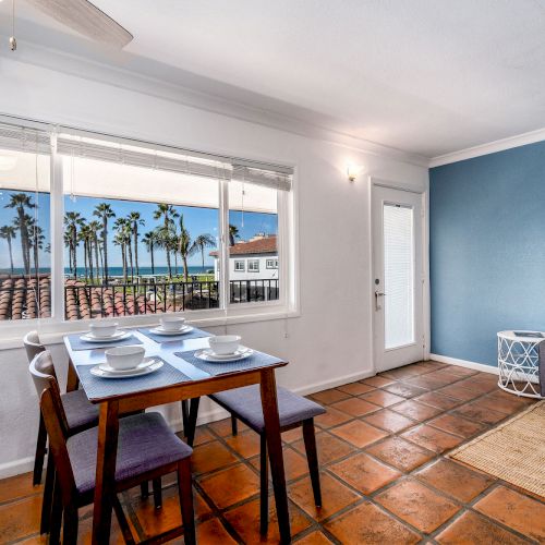 The image shows a dining area with a window view of palm trees and rooftops, a table set for four, and a small living space with a sofa and side table.