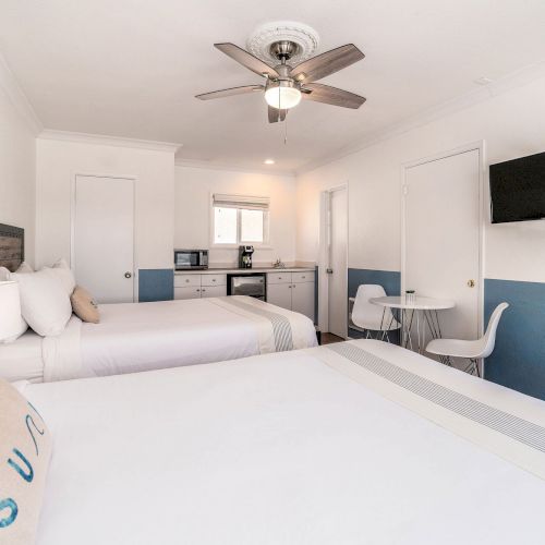 A bright, clean bedroom with two beds, a ceiling fan, wall-mounted TV, chairs, a table, a kitchenette, and a minimalist coastal decor theme ending the sentence.