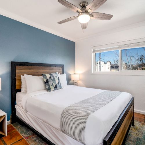 A cozy bedroom with a bed against a blue accent wall, two side tables, lamps, a ceiling fan, and a window with a view of the outside.