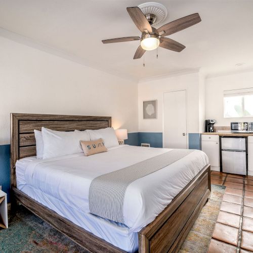 The image shows a well-lit bedroom with a large bed, ceiling fan, nightstands with lamps, and a kitchenette area with a coffee maker and mini-fridge.