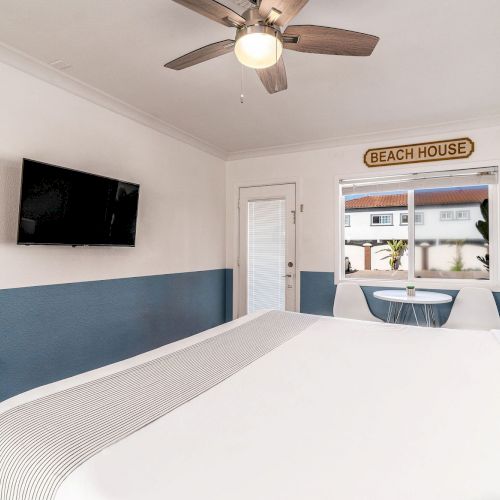 The image shows a cozy beach-themed bedroom with a ceiling fan, wall-mounted TV, door, window, and a 