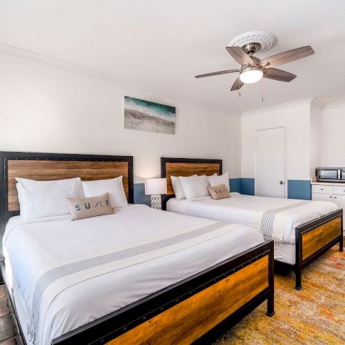 A bright, modern bedroom with two double beds, a ceiling fan, bedside tables, a kitchenette, and a painting on the wall.