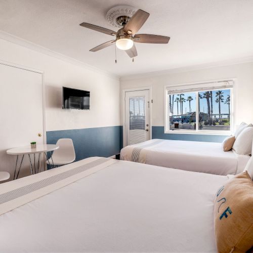 The image shows a hotel room with two beds, a small round table with two chairs, a wall-mounted TV, a ceiling fan, and a window with a beach view.