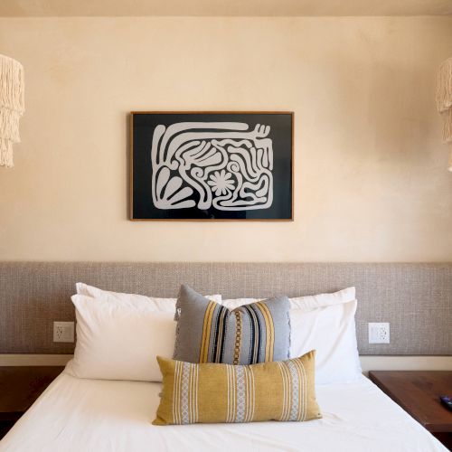 A neatly made bed between wooden side tables, with two patterned pillows, hanging lamps, and a framed abstract artwork above it.