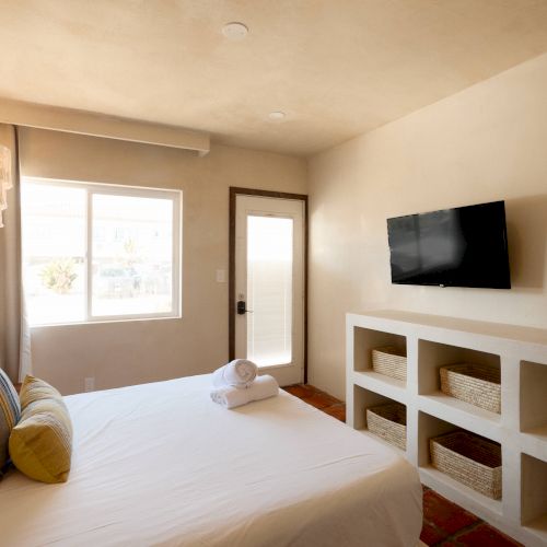 A minimalist bedroom with a bed, pillows, a wall-mounted TV, a window, a door, and built-in shelves holding baskets and towels ends the sentence.