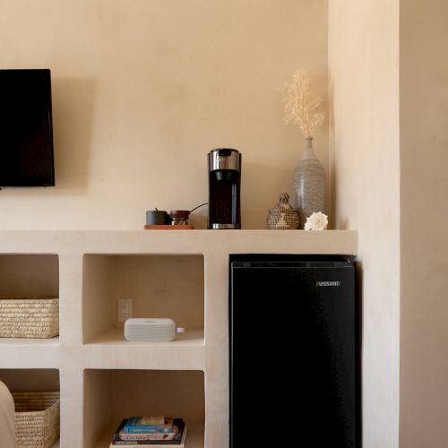 A minimalist room with built-in shelving, a coffee maker on a countertop, a small black refrigerator, and a wall-mounted TV.