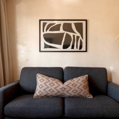 A gray sofa with a patterned pillow sits under an abstract black-and-white wall art, inside a cozy room with beige walls and natural light.