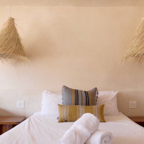 A cozy bedroom with a neatly made bed, decorative pillows, two rolled white towels, and unique woven light fixtures hanging overhead.