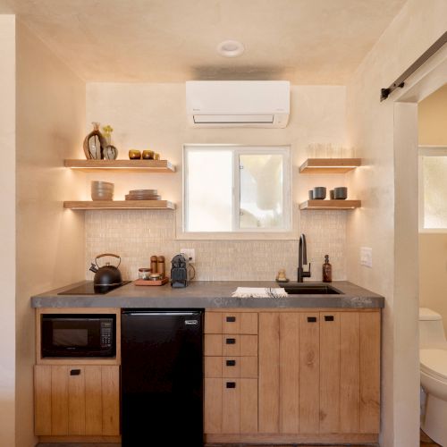 A small kitchen area with wooden cabinets, a black sink, and appliances. Wooden shelves hold dishes. A sliding door leads to a toilet.