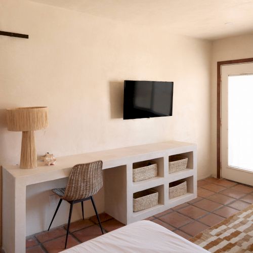 The image shows a cozy room with a desk, chair, baskets, lamp, wall-mounted TV, a door, and part of a bed, having a rustic and minimalistic design.