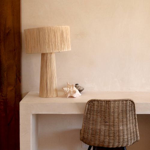 The image shows a minimalist workspace with a wooden lamp, a few decorative items on a white table, and a wicker chair with black legs.