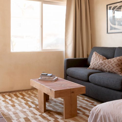 A cozy living area with a couch, a wooden coffee table, a patterned rug, and a window with curtains. There's a bed partially visible.