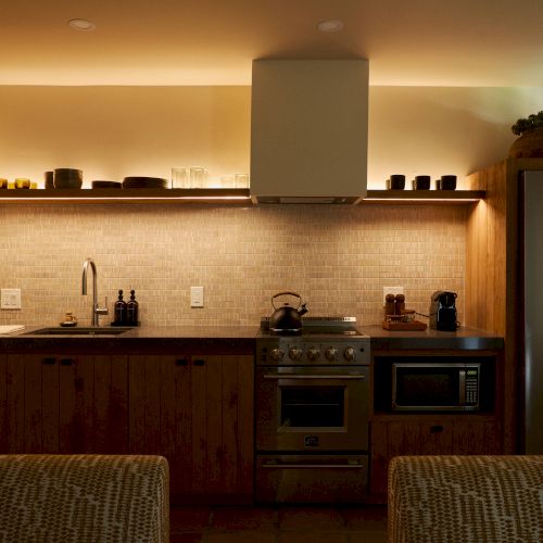 A modern kitchen with under-cabinet lighting, stove, microwave, sink, refrigerator, and minimalist décor on the shelves above the counter.