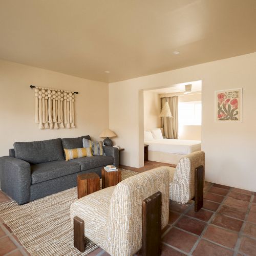 This image shows a cozy living area with a grey sofa, two cushioned chairs, and a small coffee table, adjacent to a bedroom. Decorative art adorns the walls.