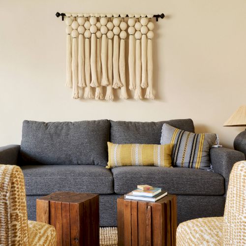 A cozy living room with a gray sofa, yellow and gray cushions, wooden coffee tables, woven chairs, a lamp, and wall hanging décor.