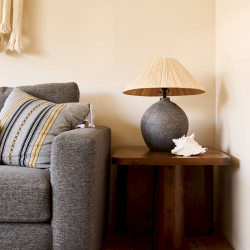 A cozy living room corner features a gray sofa with striped cushions, a wooden table with a lamp, and decorative seashells on the table.