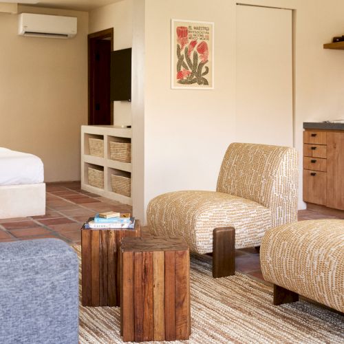 A cozy living room with two chairs, a small wooden table, patterned rug, wall art, and a view into a bedroom with a bed and a TV.