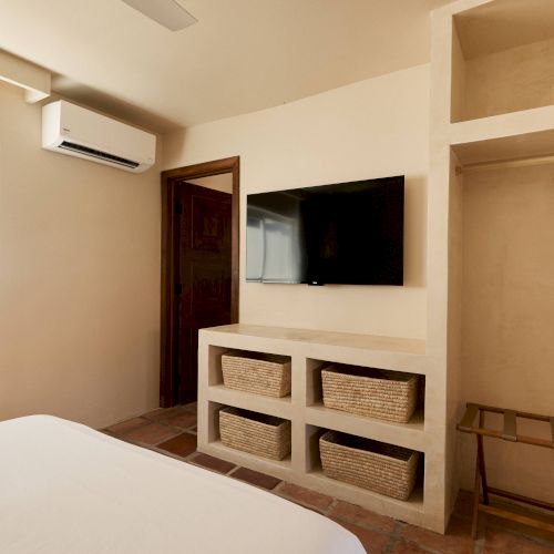 A minimalist bedroom with a bed, wall-mounted TV, air conditioner, wicker drawers, and an open closet space with shelves and a luggage rack.