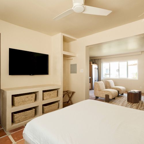 A bedroom with a bed, TV, ceiling fan, and a connected bathroom. A sitting area with chairs and a table is visible through an open doorway.