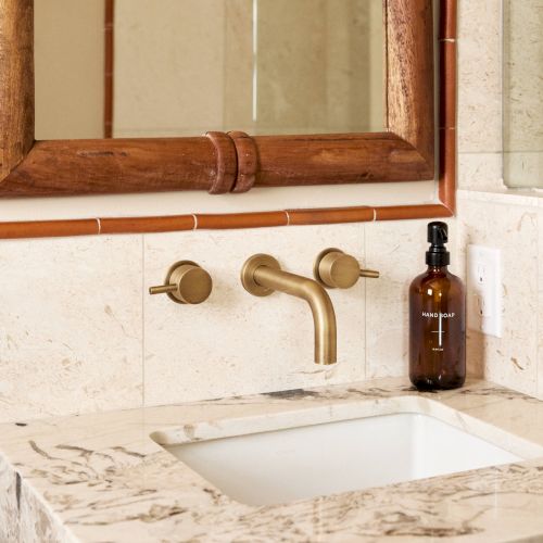 A bathroom sink with a marble countertop, brass faucet, and a wooden-framed mirror. A brown bottle is placed next to the faucet.